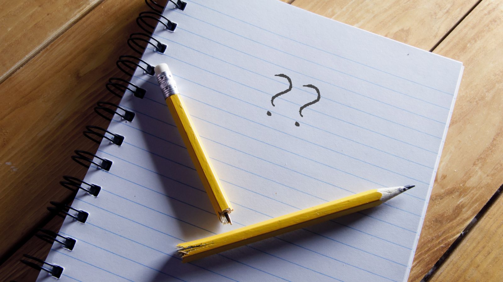 Auf einem Tisch liegt ein aufgeschlagener linierter Block mit zwei Fragezeichen in Bleistift.