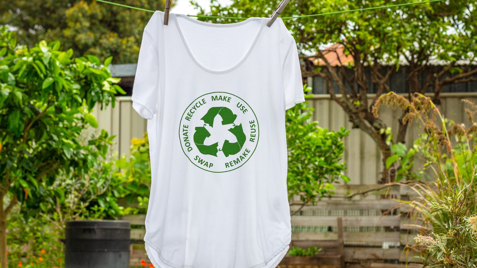 Ein weißes T-Shirt mit einer grünen Abbildung eines Recyclingkreislaufs hängt auf einer Leine in einem grünen Garten