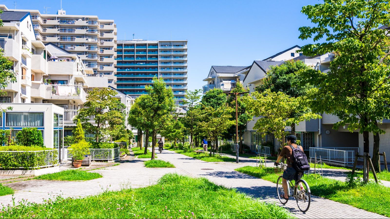 Ein Mann fährt auf dem Fahrrad durch eine begrünte Wohnsiedlung einer Stadt. Im Hintergrund sind hohe Gebäude zu sehen.