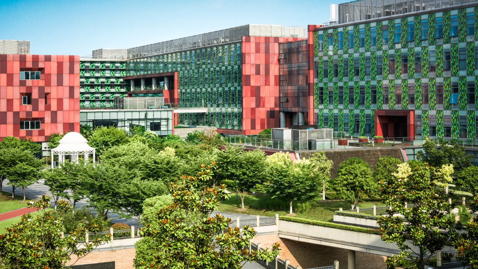 Von Bürogebäuden eingerahmte Grünfläche mit Bäumen, Wegen und einem Pavillon. Im Hintergrund ist blauer Himmel zu sehen.