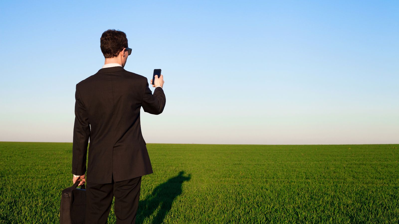 Ein junger Geschäftsmann mit  Sonnenbrille und Aktentasche richtet sein Smartphone in Richtung einer endlos weiten Grasebene, welche sich bis zum Horizont erstreckt. Der Himmel ist wolkenlos.