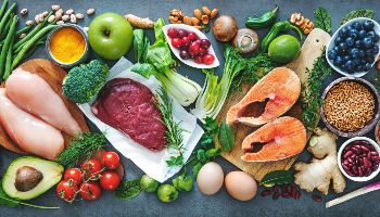 Eine dunkle Arbeitsfläche ist flächendeckend mit diversen frischen Lebensmitteln bedeckt.  Die Lebensmittelauswahl erstreckt sich von Obst und Gemüse neben Fleisch, Fisch und Eiern.