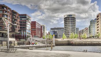 Der Blick fällt auf eine moderne Stadt über ein Wasser mit Bänken am gestalteten Ufer. 