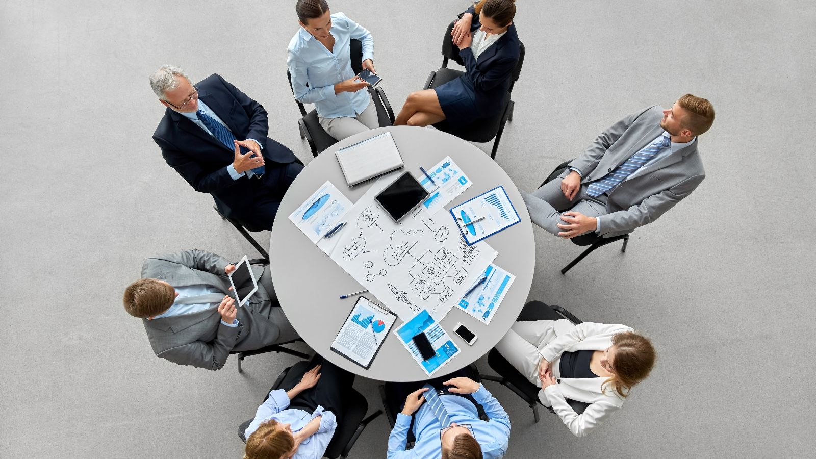 Vogelperspektive auf eine Gruppe von Menschen in formalen Anzügen an einem runden Tisch sitzend. Darauf sind Planungsmaterialien erkennbar. Der Hintergrund ist ein grauer Boden.