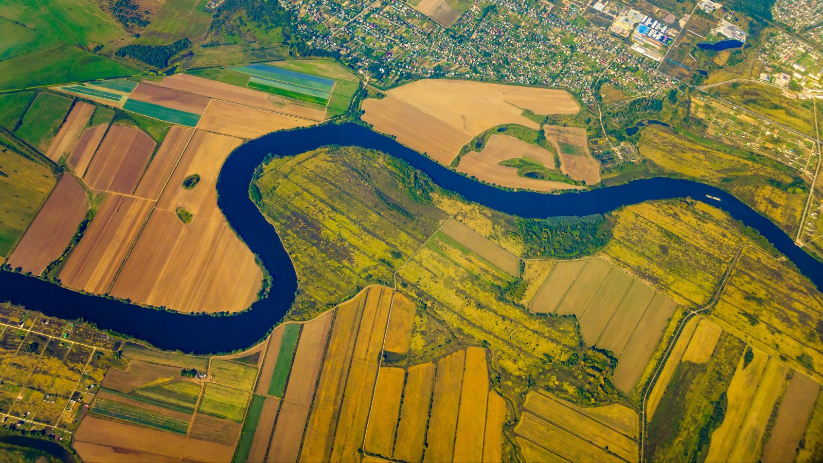 Vogelperspektive auf einen Fluss, welcher durch landwirtschaftlich genutzte Flächen mäandert. Am oberen Rand ist ein Siedlungsgebiet zu sehen.