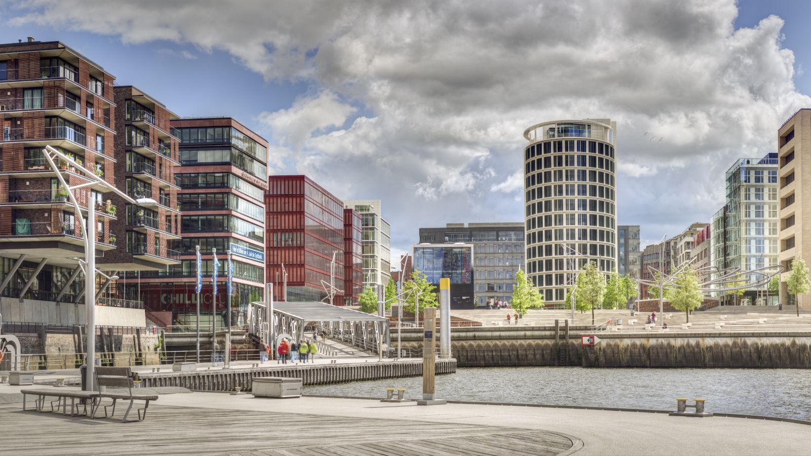 Der Blick fällt auf eine moderne Stadt über ein Wasser mit Bänken am gestalteten Ufer.