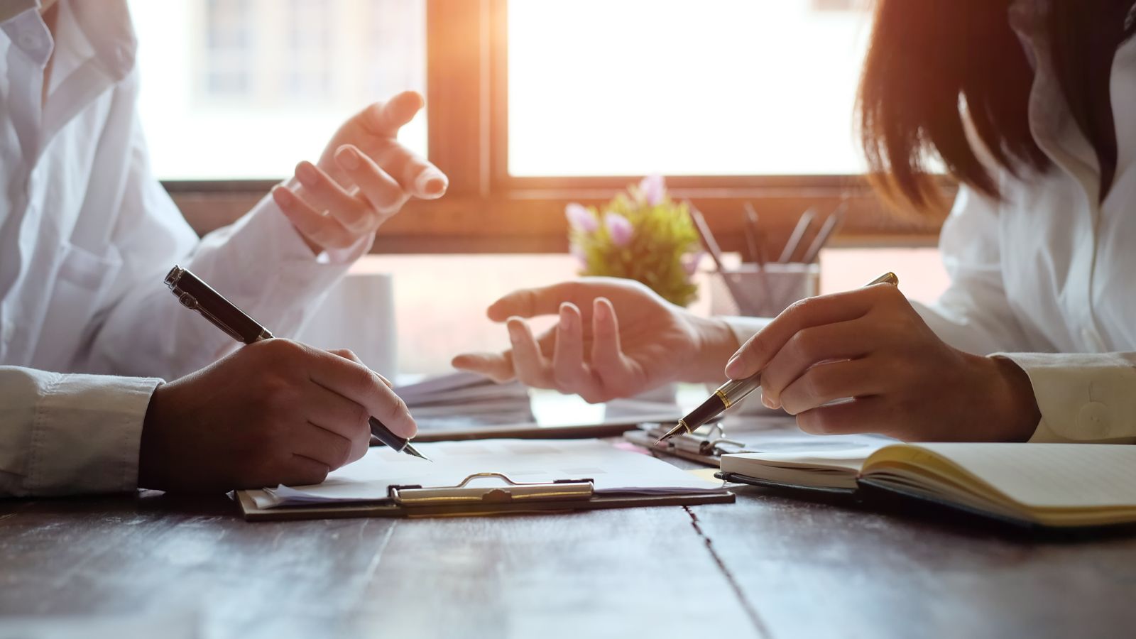 Zwei Personen sitzen an einem Holztisch und halten je eine Hand mit Stift über ein Papier.