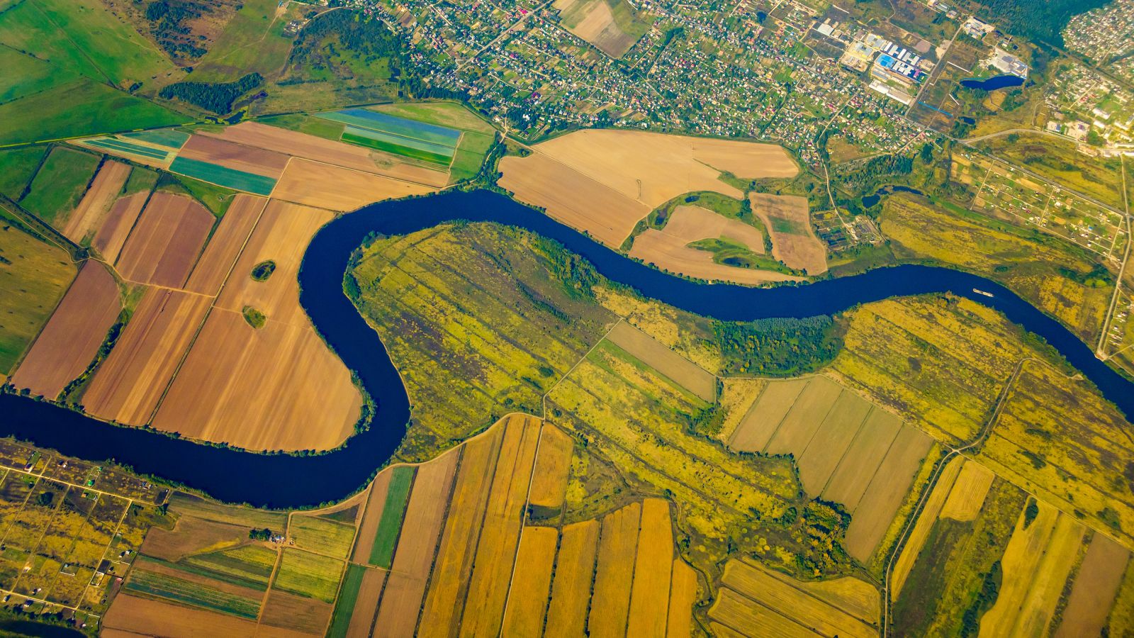 Vogelperspektive auf einen Fluss, welcher durch landwirtschaftlich genutzte Flächen mäandert. Am oberen Rand ist ein Siedlungsgebiet zu sehen.  