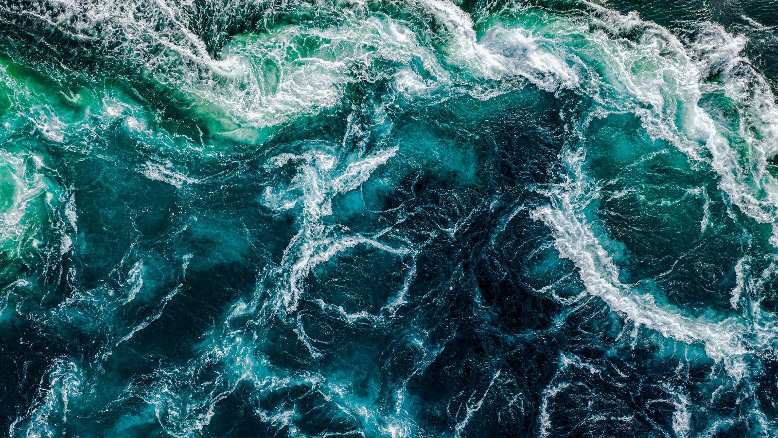 Aus der Vogelperspektive fällt der Blick auf das dunkelblaue Wasser eines aufgewühlten Meeres. Die Gischt bildet ein weißes Muster auf der Wasseroberfläche.