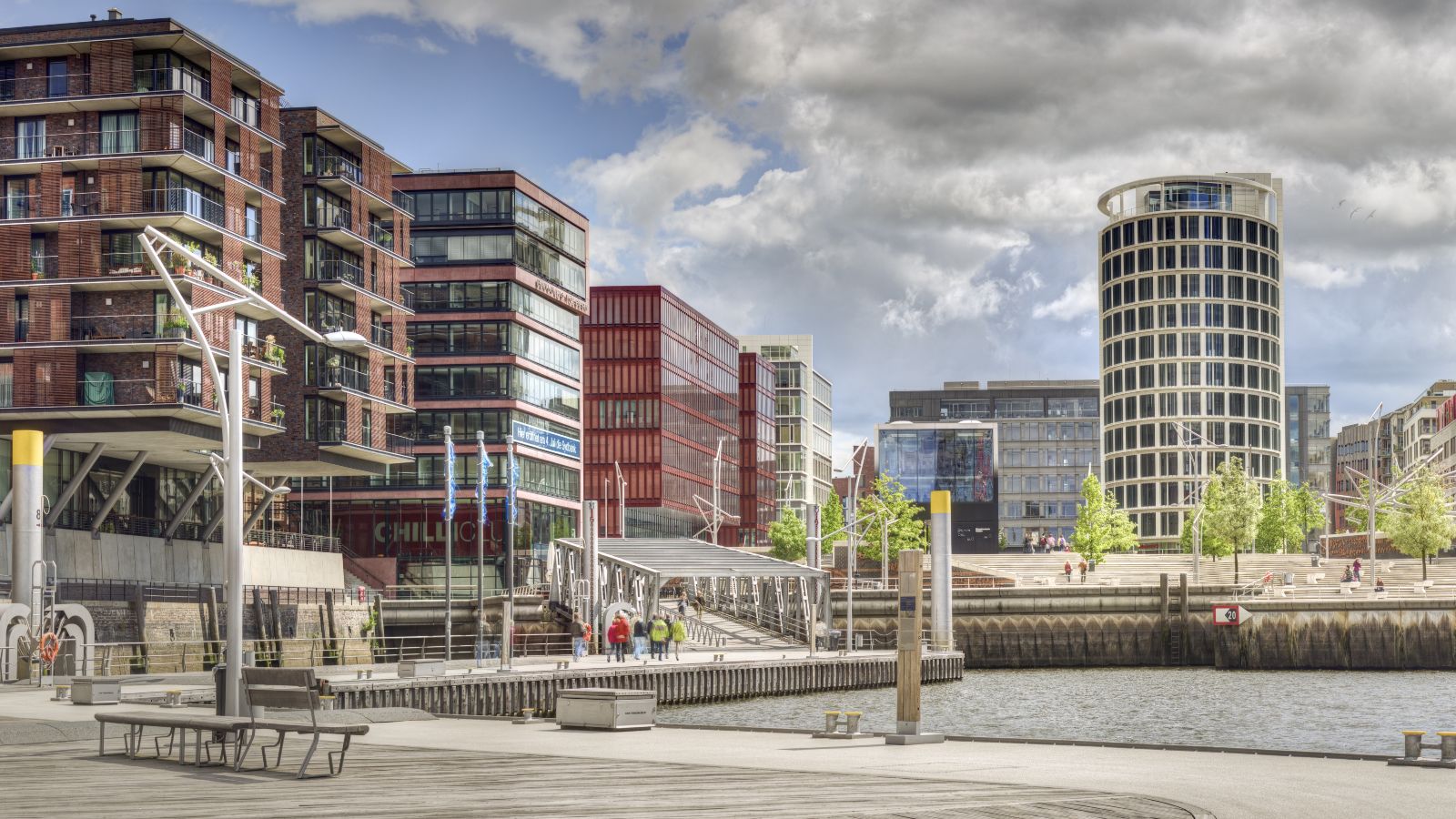 Der Blick fällt auf eine moderne Stadt über ein Wasser mit Bänken am gestalteten Ufer.