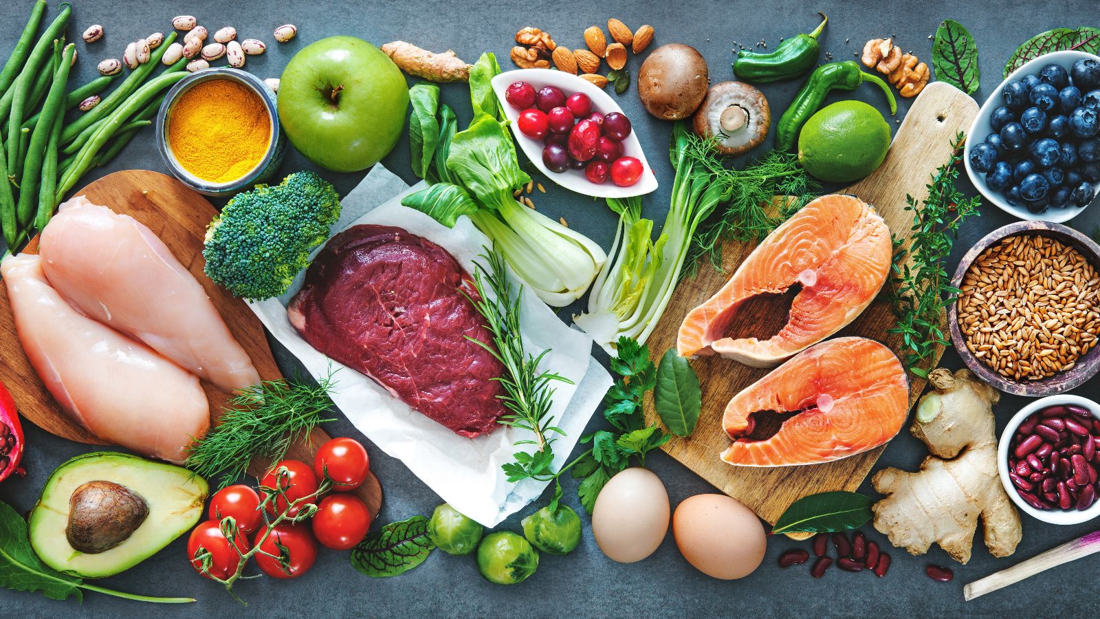 Eine dunkle Arbeitsfläche ist flächendeckend mit diversen frischen Lebensmitteln bedeckt. Die Lebensmittelauswahl erstreckt sich von Obst und Gemüse neben Fleisch, Fisch und Eiern.