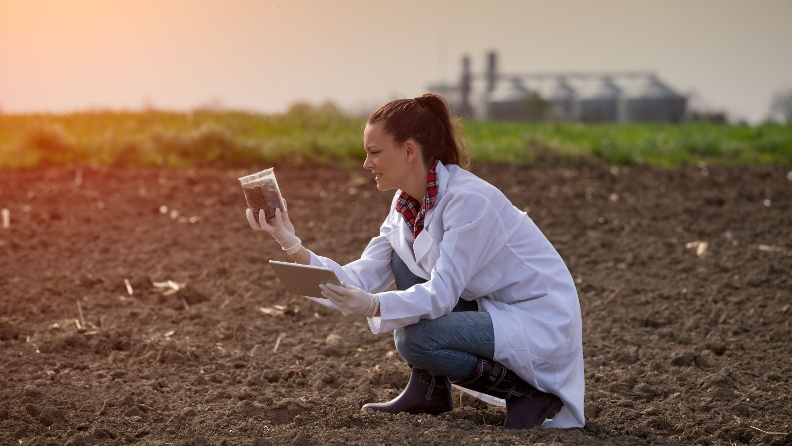 Eine Frau mit weißem Laborkittel kniet auf einem Acker und hält ein Becherglas mit einer Bodenprobe und ein Tablet in den Händen. Im Hintergrund sind eine Wiese und eine Biogasanlage zu sehen.