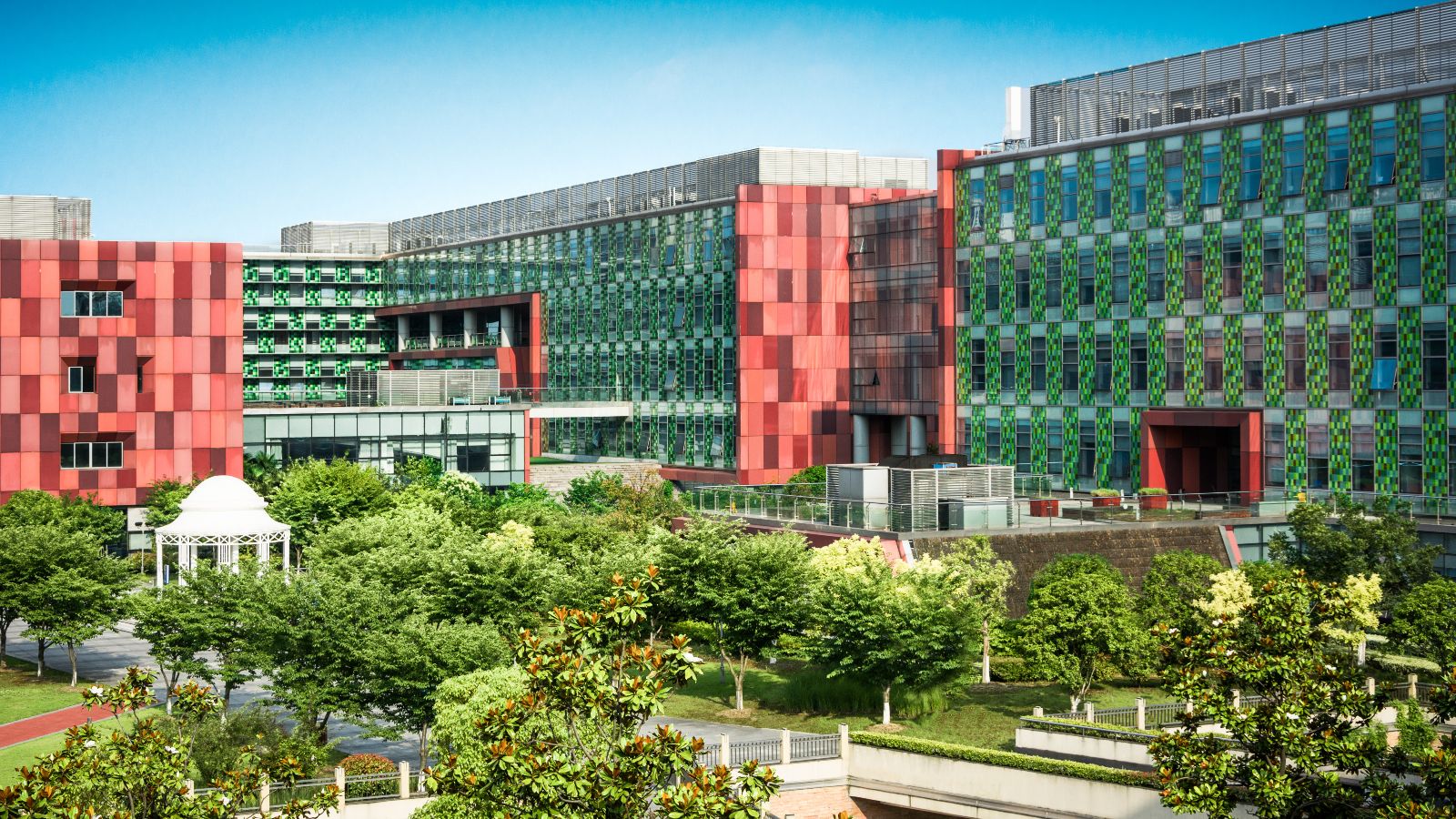 Von Bürogebäuden eingerahmte Grünfläche mit Bäumen, Wegen und einem Pavillon. Im Hintergrund ist blauer Himmel zu sehen.