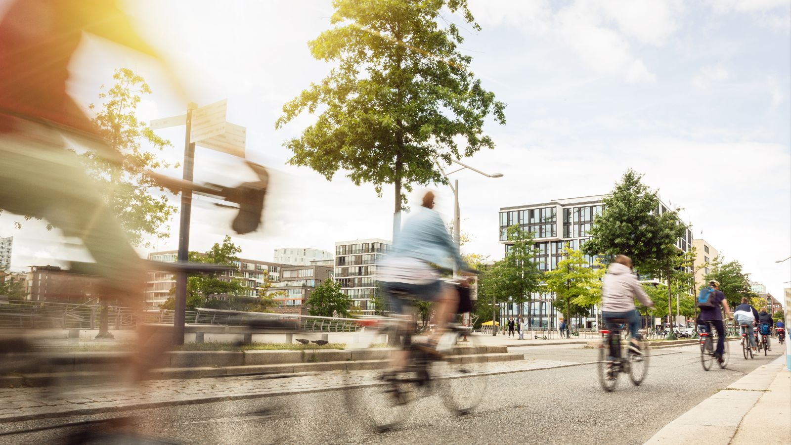Mehrere Fahrradfahrer fahren hintereinander auf einem Fahrradweg in einer Stadt. Im Hintergrund sind Häuser und Bäume zu sehen.
