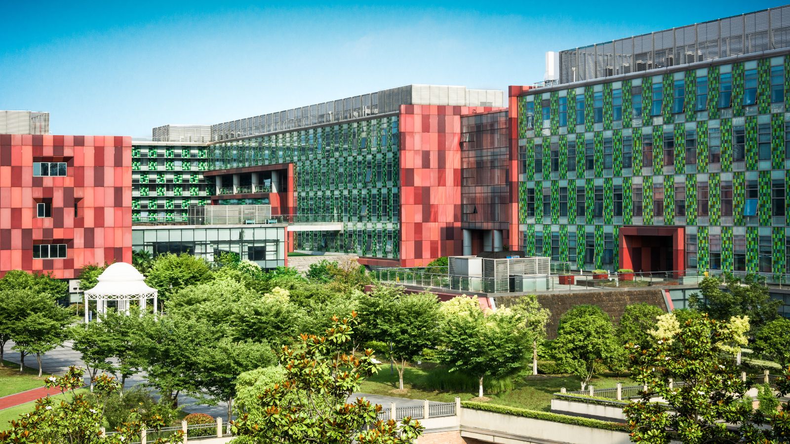 Von Bürogebäuden eingerahmte Grünfläche mit Bäumen, Wegen und einem Pavillon. Im Hintergrund ist blauer Himmel zu sehen.