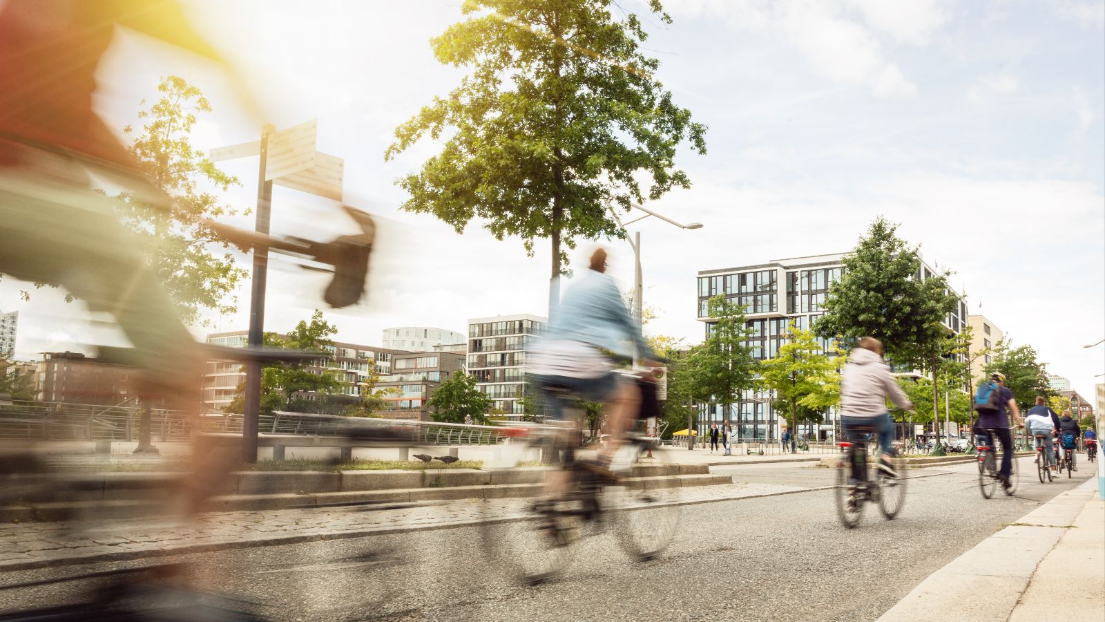 Mehrere Fahrradfahrer fahren hintereinander auf einem Fahrradweg in einer Stadt. Im Hintergrund sind Häuser und Bäume zu sehen.