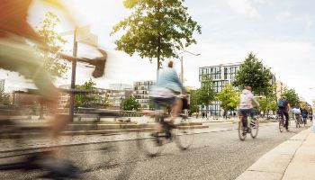 Mehrere Fahrradfahrer fahren hintereinander auf einem Fahrradweg in einer Stadt. Im Hintergrund sind Häuser und Bäume zu sehen.