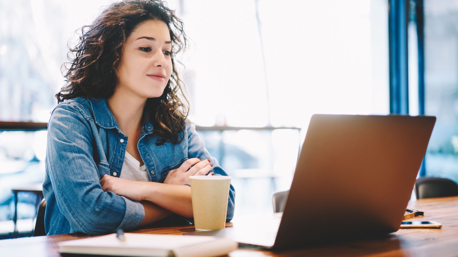 Eine junge Frau sitzt aufmerksam mit Kaffeebecher und Notizblock vor einem aufgeklappten Laptop. 