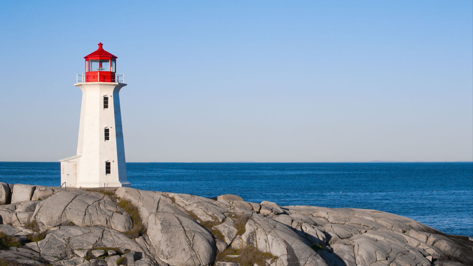 Ein weißer Leuchtturm mit rotem Dach steht auf einem Felsen vor blauem Meer mit blauem Himmel.