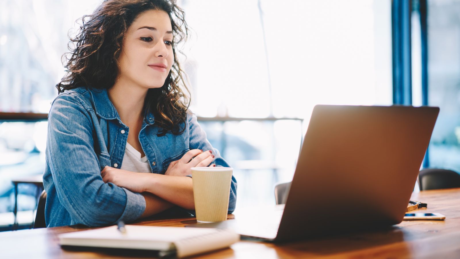 Eine junge Frau sitzt aufmerksam mit Kaffeebecher und Notizblock vor einem aufgeklappten Laptop. 