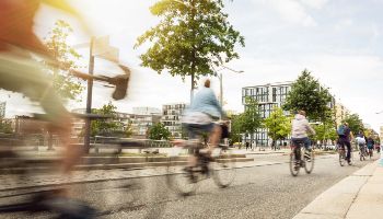 Mehrere Fahrradfahrer fahren hintereinander auf einem Fahrradweg in einer Stadt. Im Hintergrund sind Häuser und Bäume zu sehen.