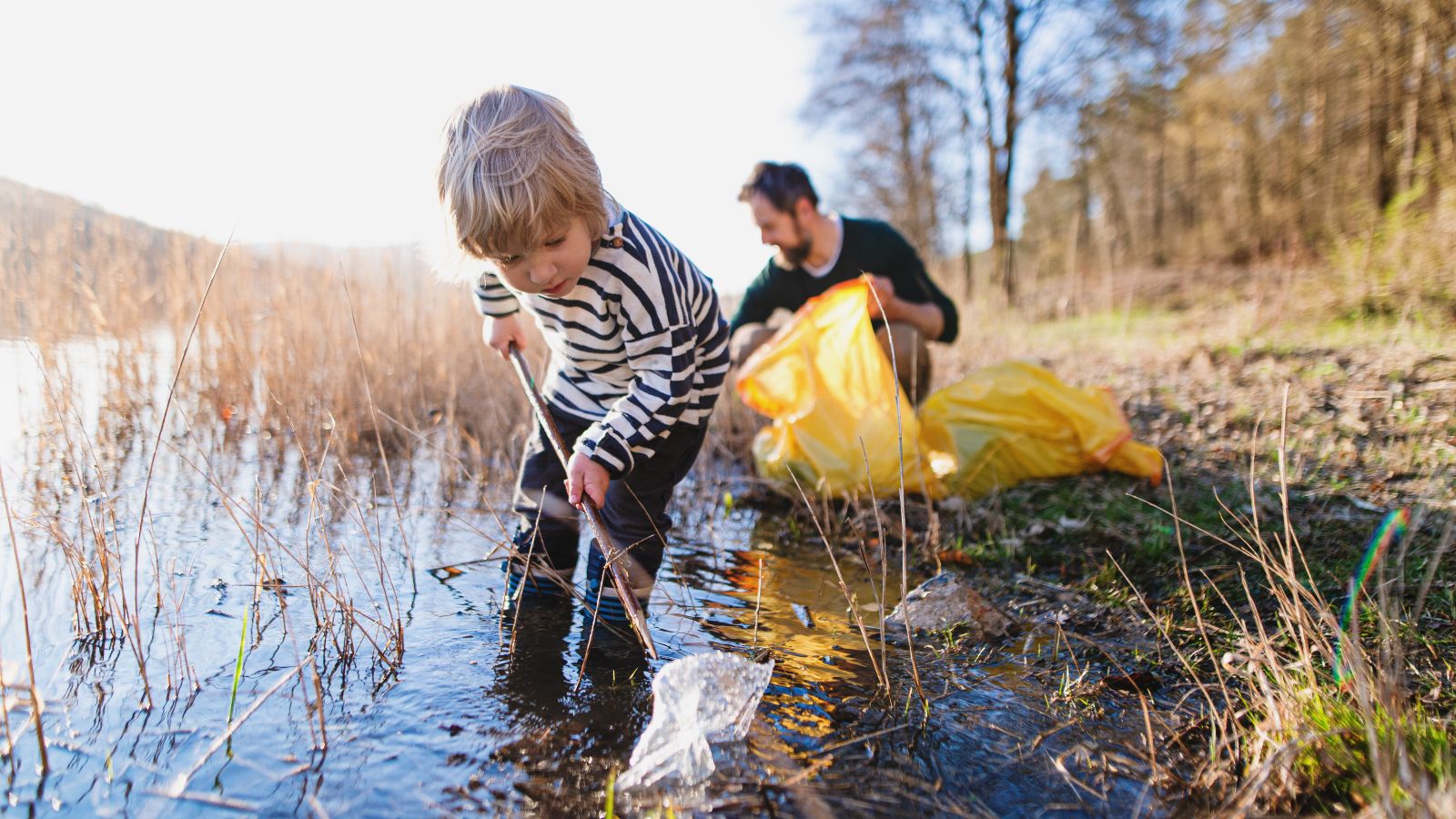 Kind und erwachsener Mann sammeln Müll aus seichtem Gewässer am Uferrand eines Sees, bei strahlender Sonne.