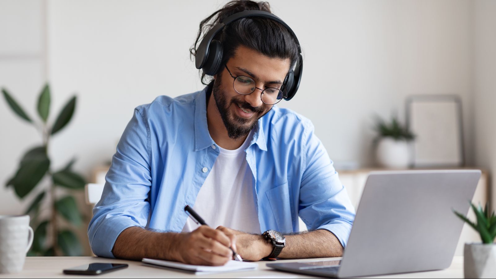Ein Mann sitzt mit Kopfhörern an einem Schreibtisch vor einem Laptop und schreibt etwas auf einen Notizblock. Im Hintergrund sind unscharf Möbel und Pflanzen zu sehen.