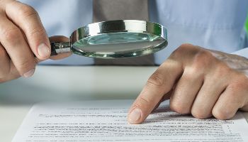 There is a document on a table. Above it are two hands. One hand holds a magnifying glass, the other points to a place in the document with its index finger.