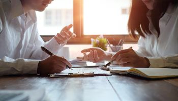 Two people sit at a wooden table, each holding a hand with a pencil over a piece of paper. 