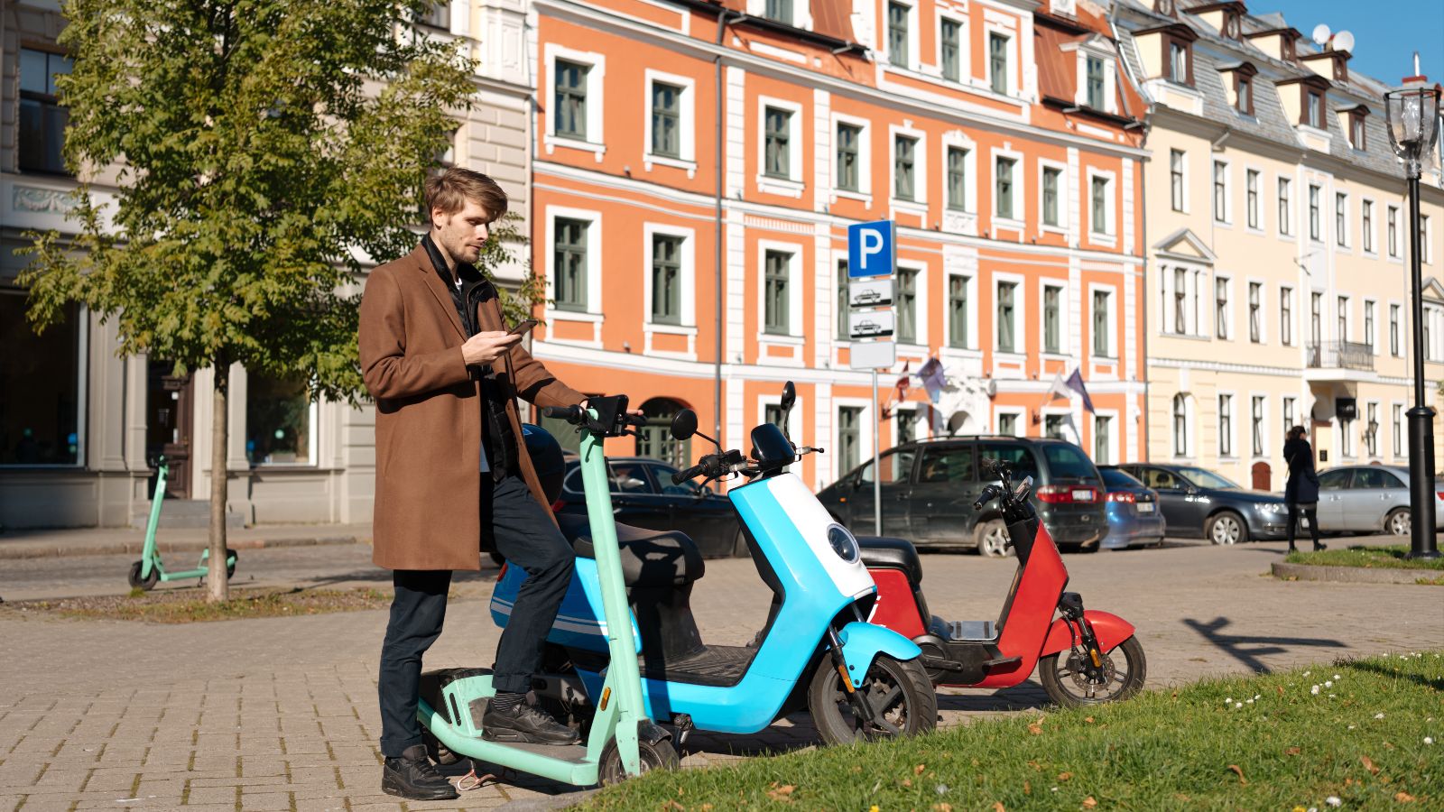 Ein junger Mann steht an einem E-Scooter und schaut auf sein Smartphone. Daneben stehen zwei E-Roller und im Hintergrund sind Häuserfassaden und Autos zu sehen.