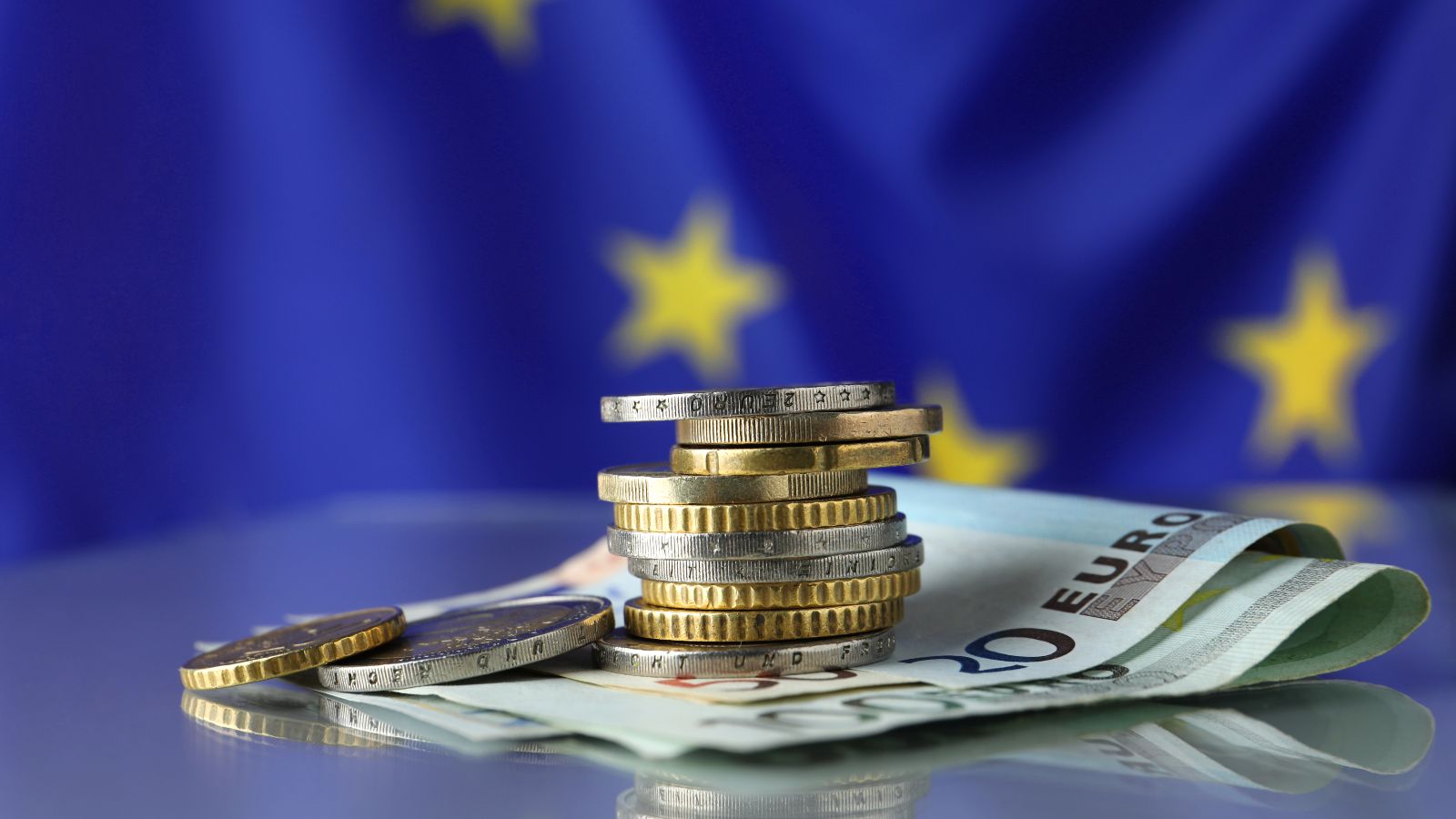 Ein Euro-Münzenstapel liegt auf Euro-Geldscheinen vor einer EU-Flagge