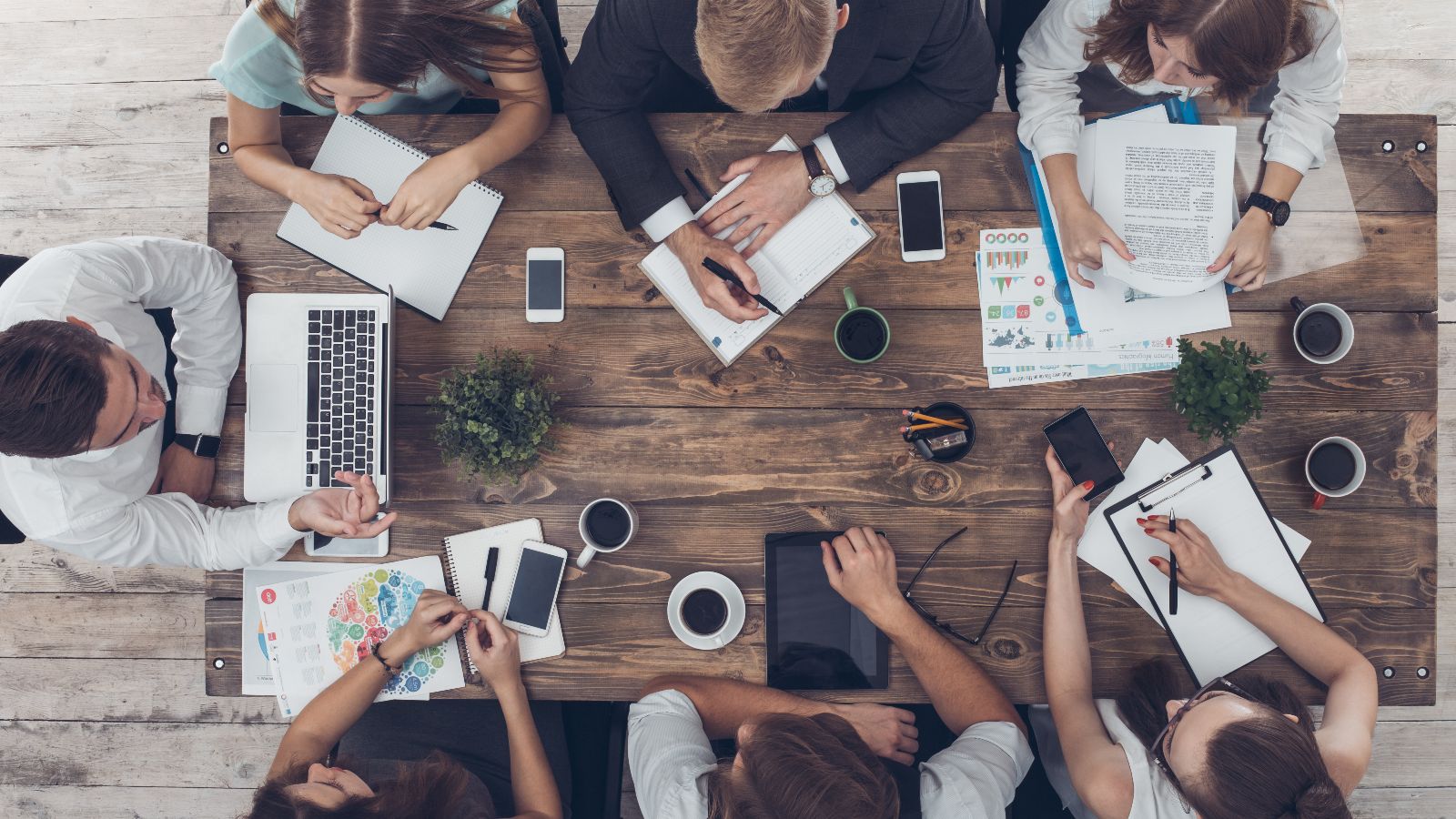 Draufsicht auf einen Tisch mit Unterlagen, Laptops, Tablets, Smartphones und Kaffeetassen. Um den Tisch sitzen sieben Personen und diskutieren.