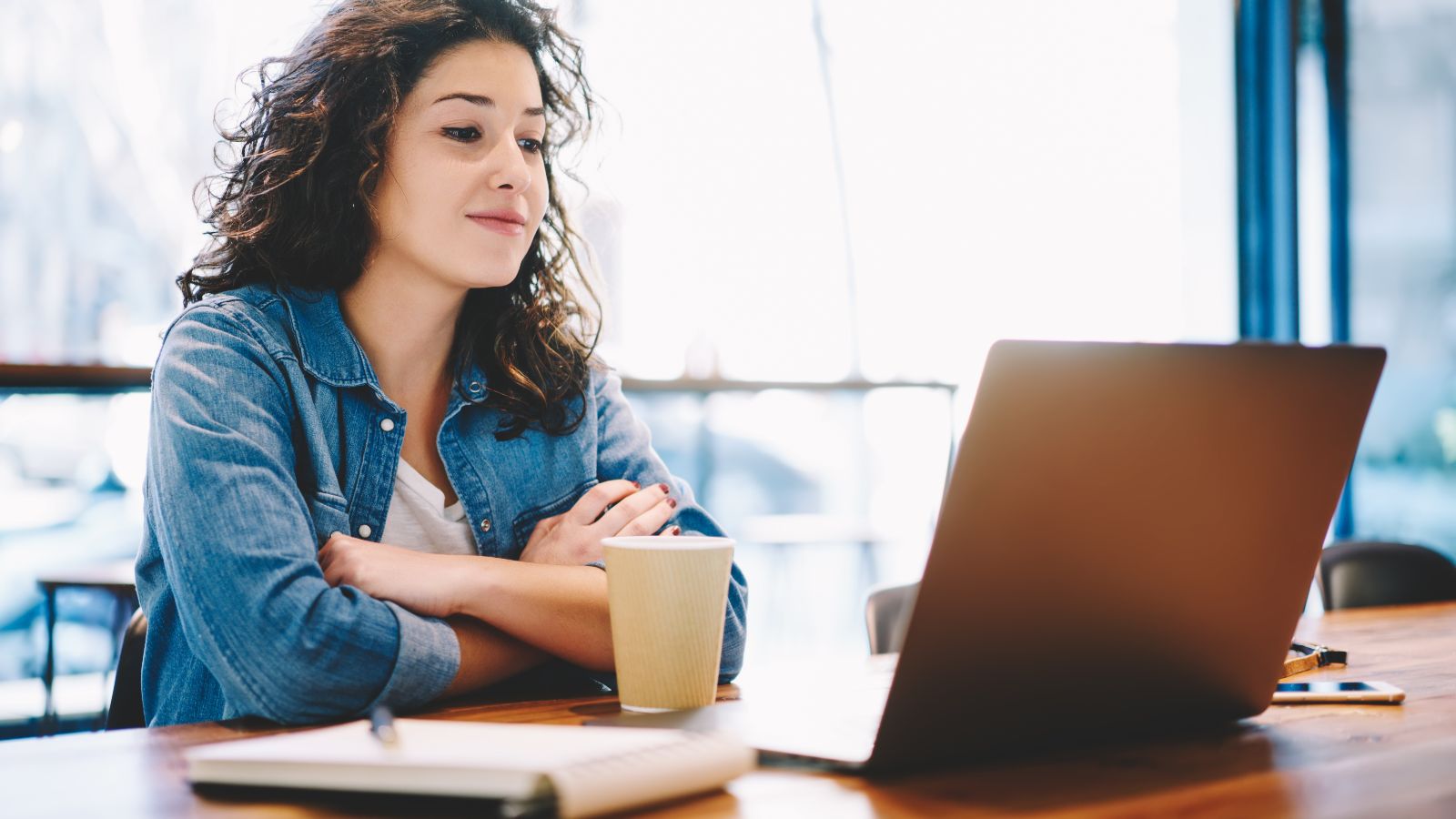 Eine junge Frau sitzt aufmerksam mit Kaffeebecher und Notizblock vor einem aufgeklappten Laptop.