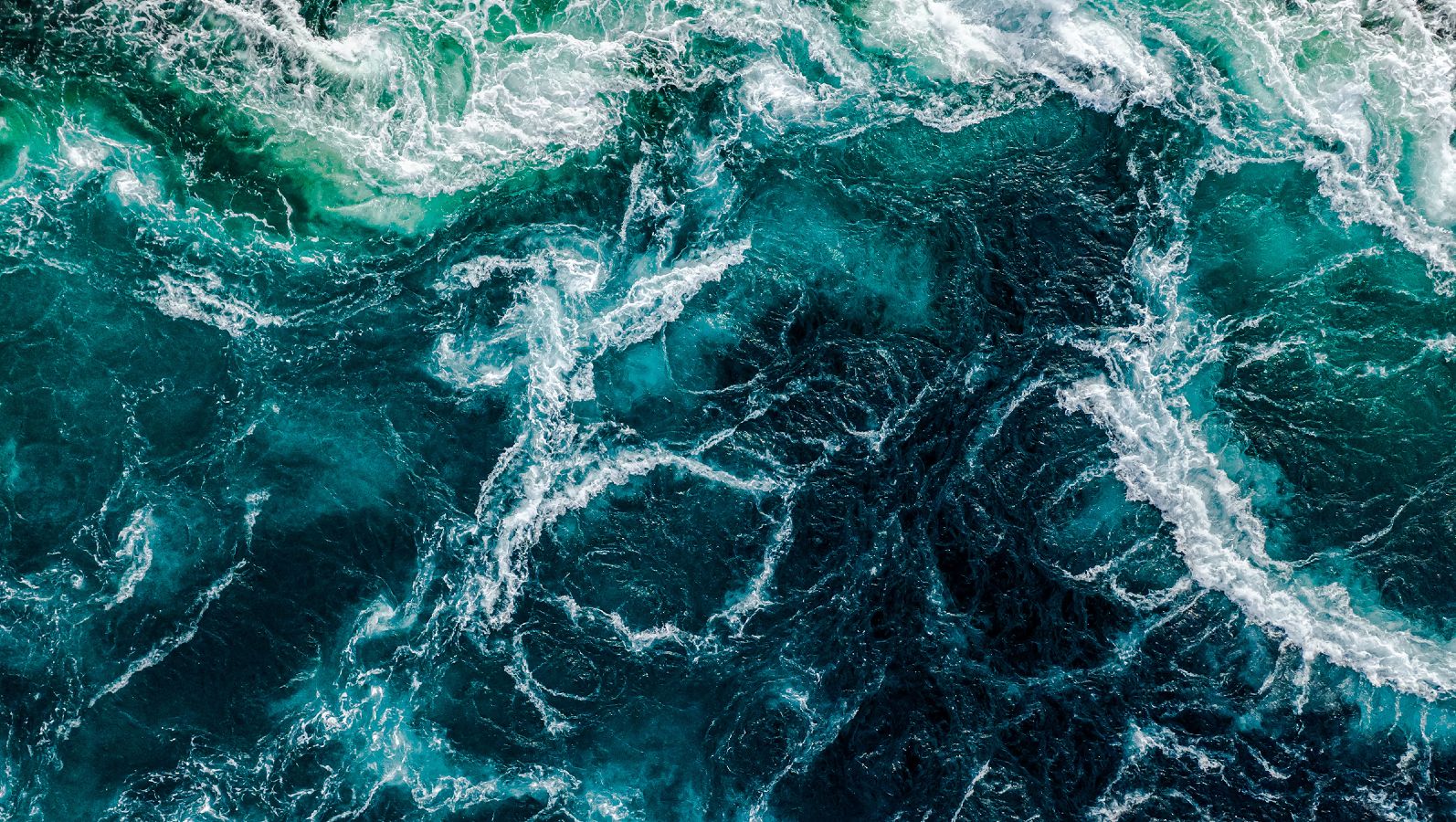 Aus der Vogelperspektive fällt der Blick auf das dunkelblaue Wasser eines aufgewühlten Meeres. Die Gischt bildet ein weißes Muster auf der Wasseroberfläche.