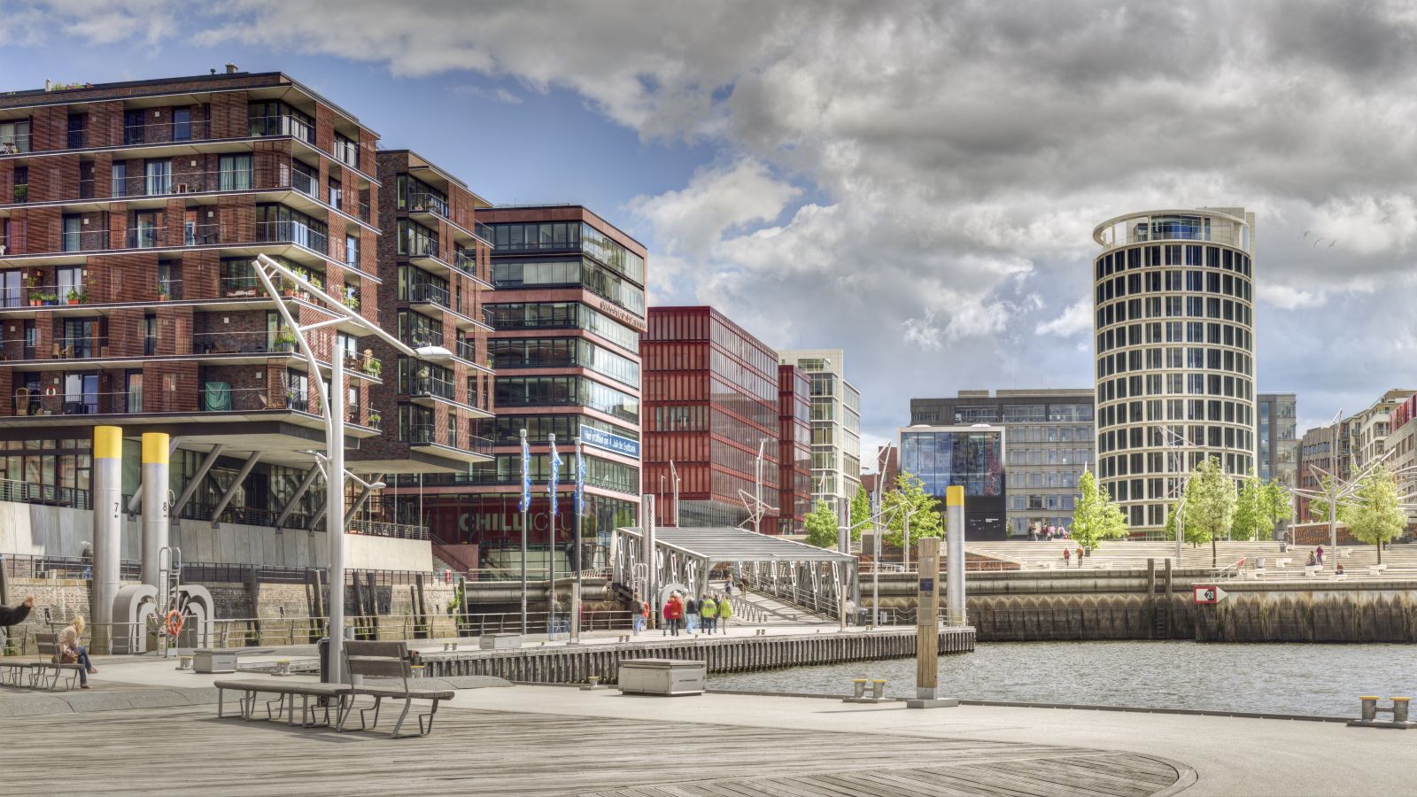 Der Blick fällt auf eine moderne Stadt über ein Wasser mit Bänken am gestalteten Ufer. 
