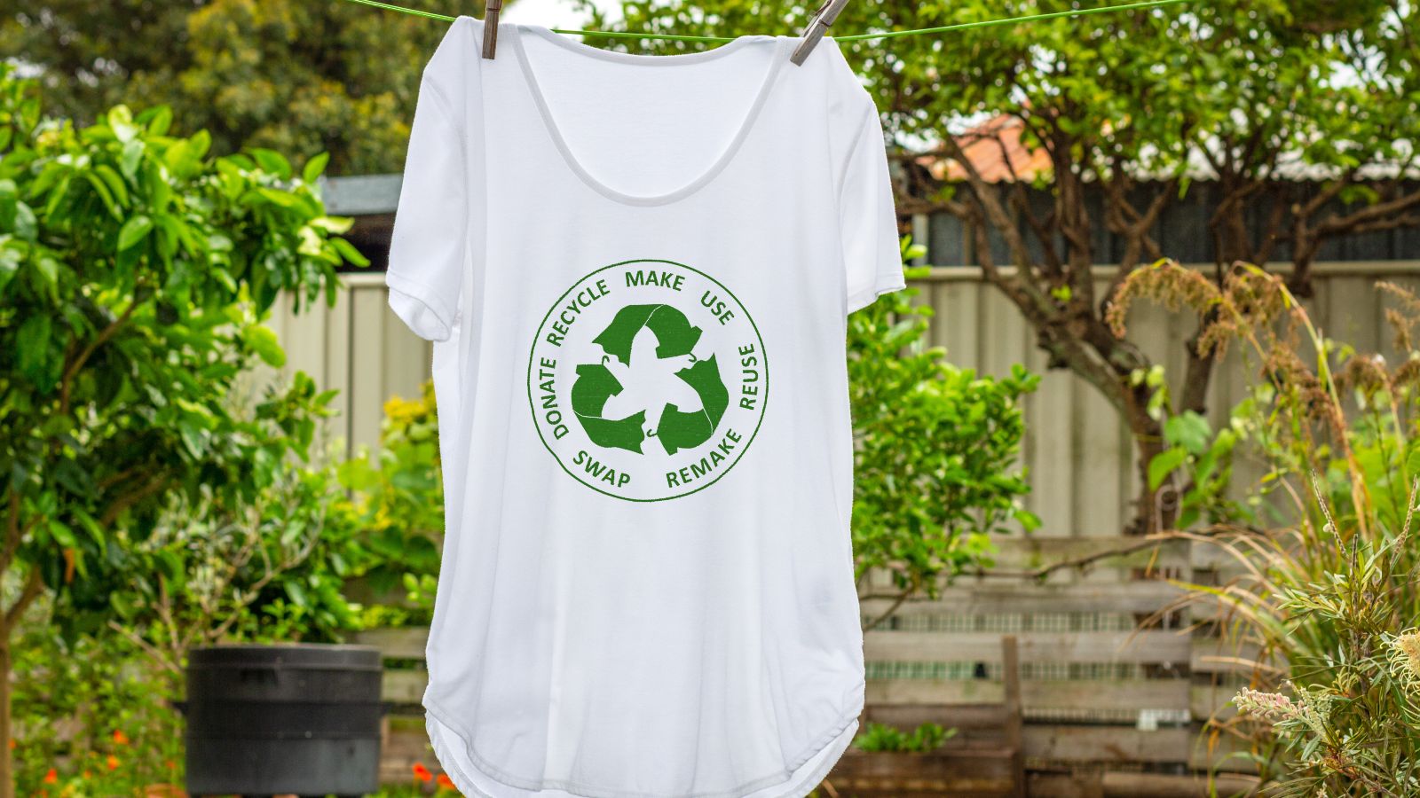 Ein weißes T-Shirt mit einer grünen Abbildung eines Recyclingkreislaufs hängt auf einer Leine in einem grünen Garten