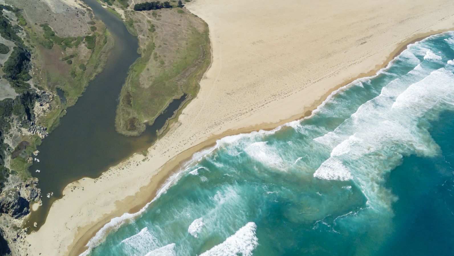 Sandiges Festland trifft an einer Küste auf türkis-blaues Meer. Große Wellen brechen in der Brandung kurz vor dem Strand.