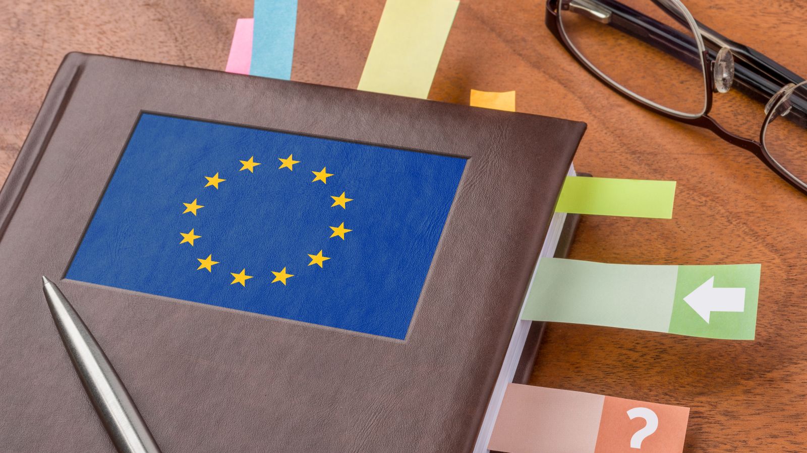 Ein Notizbuch mit einer EU-Flagge als Logo und eine schwarze Lesebrille liegen auf einem Tisch. Zwischen den Seiten im Notizbuch sind bunte Post-its eingeklebt. Auf dem Notizbuch liegt ein Stift.