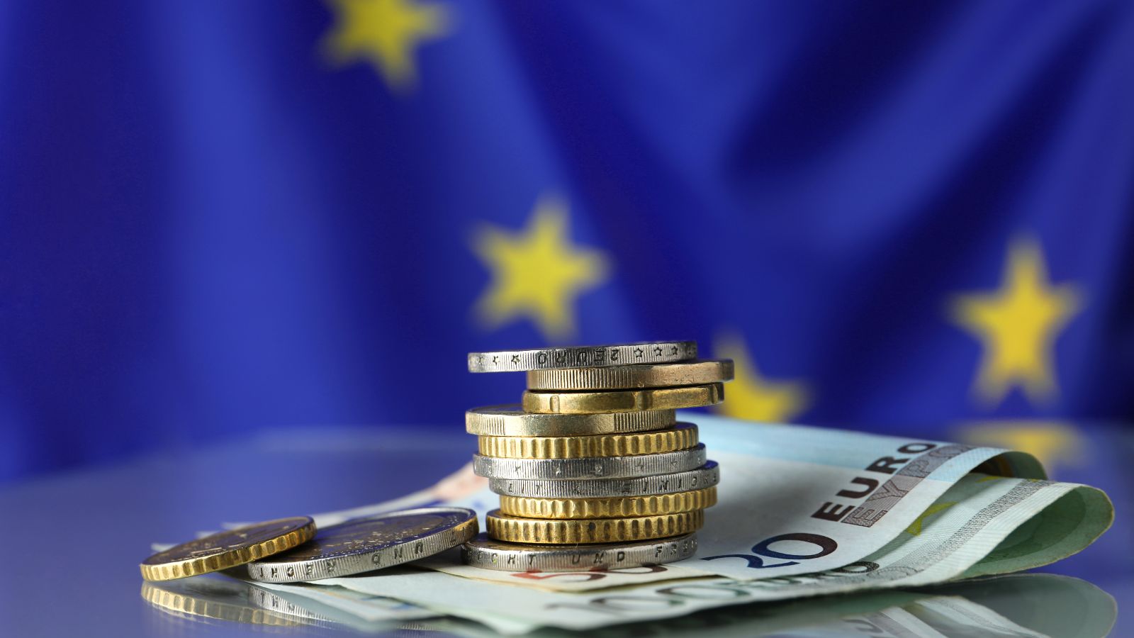 Ein Euro-Münzenstapel liegt auf Euro-Geldscheinen vor einer EU-Flagge