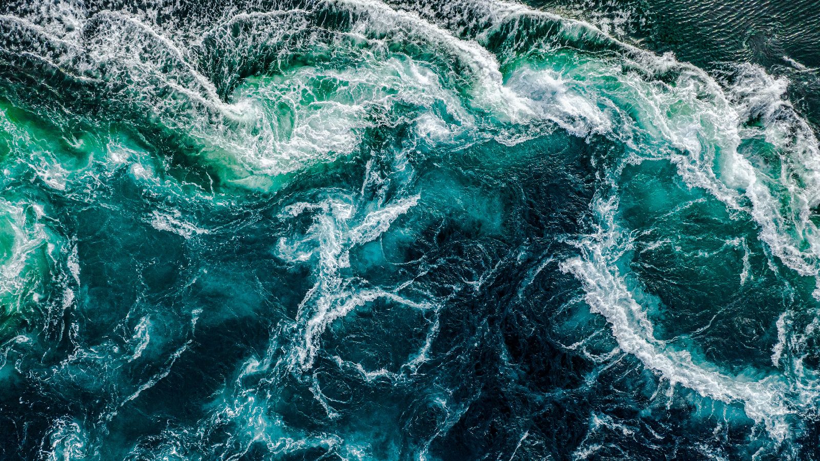 Aus der Vogelperspektive fällt der Blick auf das dunkelblaue Wasser eines aufgewühlten Meeres. Die Gischt bildet ein weißes Muster auf der Wasseroberfläche.