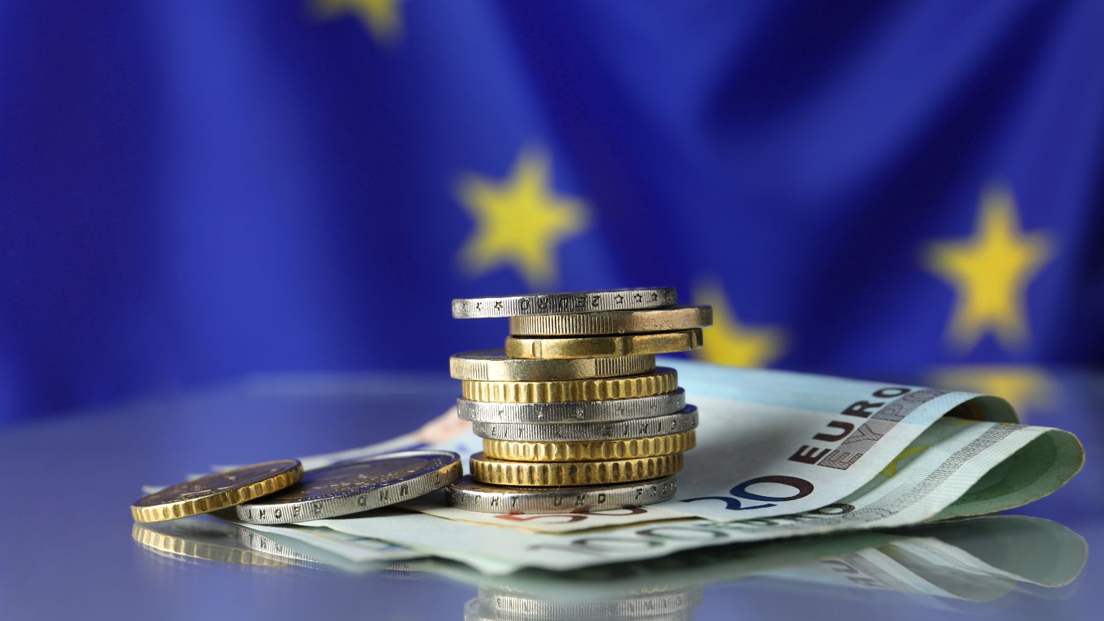 Ein Euro-Münzenstapel liegt auf Euro-Geldscheinen vor einer EU-Flagge.