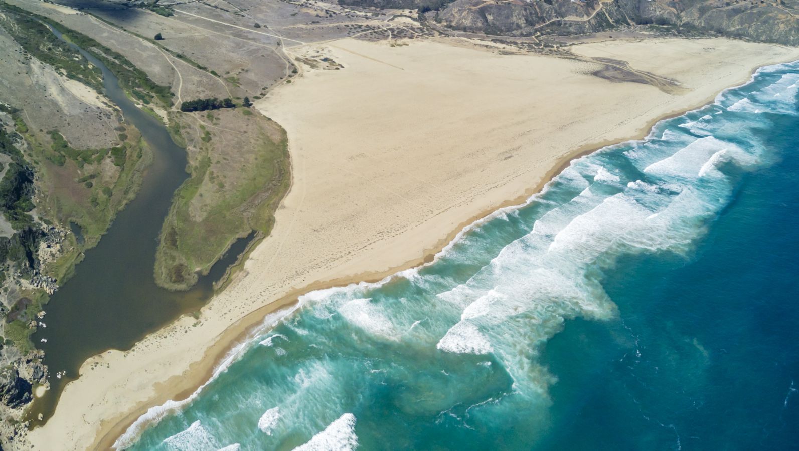 Sandiges Festland trifft an einer Küste auf türkis-blaues Meer. Große Wellen brechen in der Brandung kurz vor dem Strand.