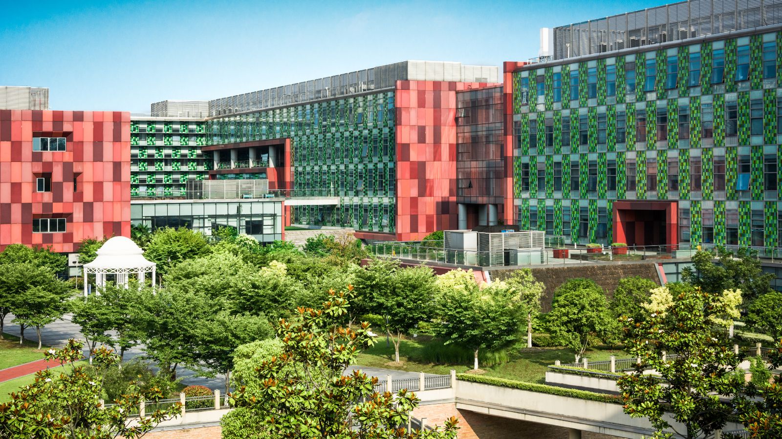 Von Bürogebäuden eingerahmte Grünfläche mit Bäumen, Wegen und einem Pavillon. Im Hintergrund ist blauer Himmel zu sehen.