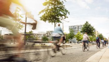 Several cyclists ride one behind the other on a bicycle path in a city. Houses and trees can be seen in the background.