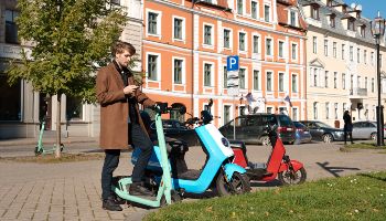 Ein junger Mann steht an einem E-Scooter und schaut auf sein Smartphone. Daneben stehen zwei E-Roller und im Hintergrund sind Häuserfassaden und Autos zu sehen.