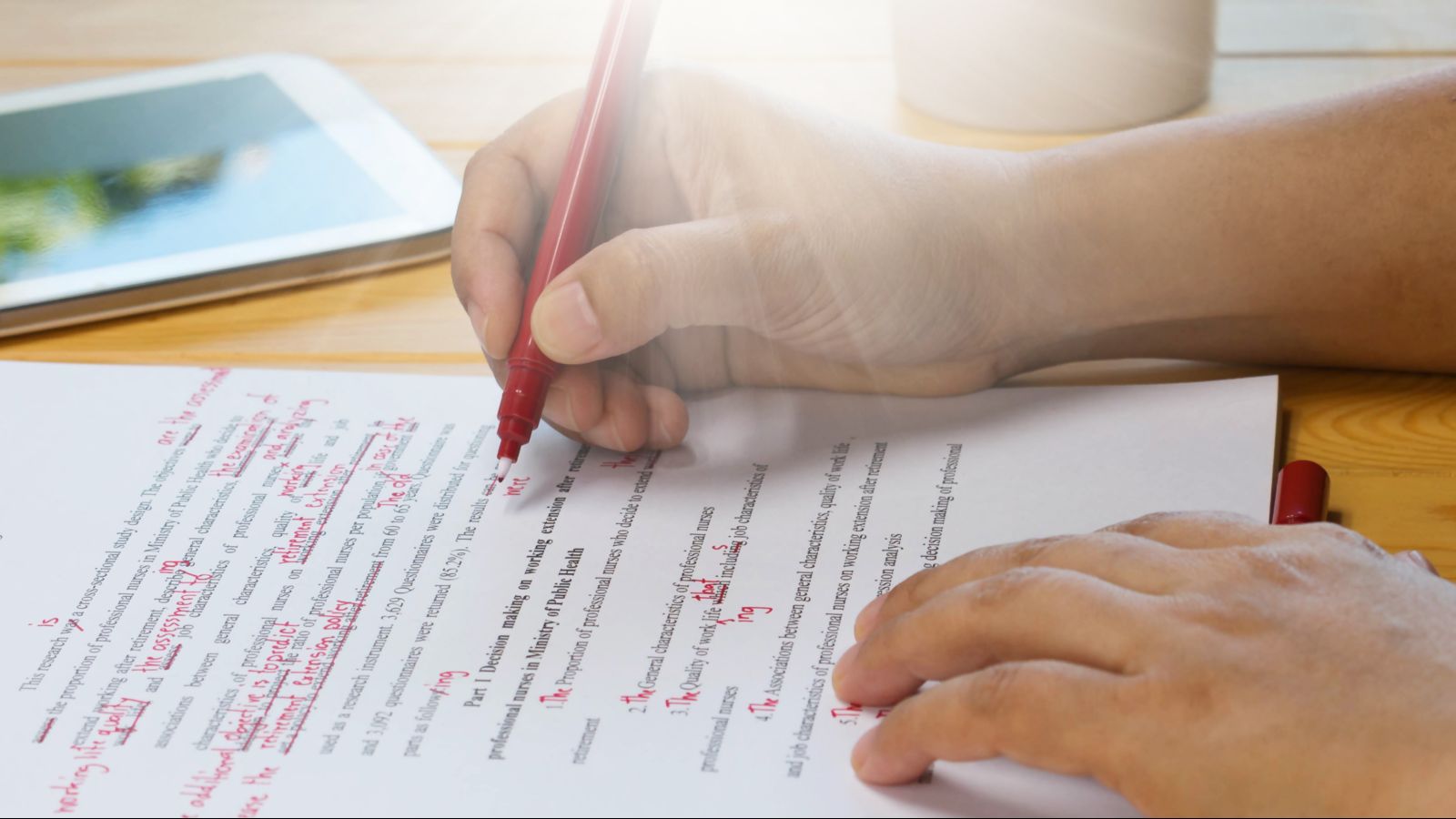 Auf einem Tisch liegt ein Dokument. Darüber sind zwei Hände. Eine Hand hält einen Rotstift und macht Korrekturen, die andere hält die Stiftkappe. Neben dem Dokument liegt ein Handy.