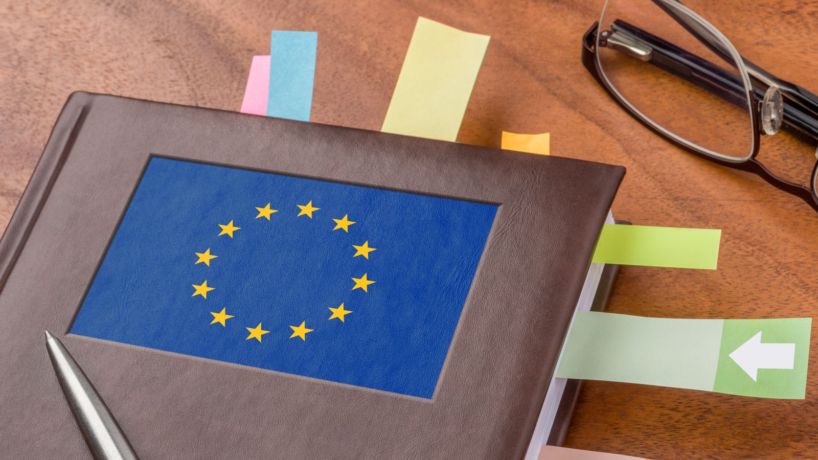 Ein Notizbuch mit einer EU-Flagge als Logo und eine schwarze Lesebrille liegen auf einem Tisch. Zwischen den Seiten im Notizbuch sind bunte Post-its eingeklebt. Auf dem Notizbuch liegt ein Stift.