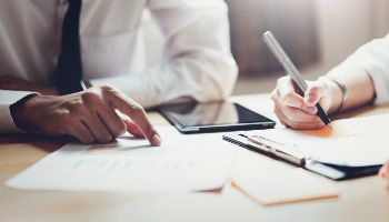 Ein Mann und eine Frau sitzen zusammen geschäftig an einem Tisch. Der Mann deutet auf eine Tabelle, während die Frau Notizen schreibt.
