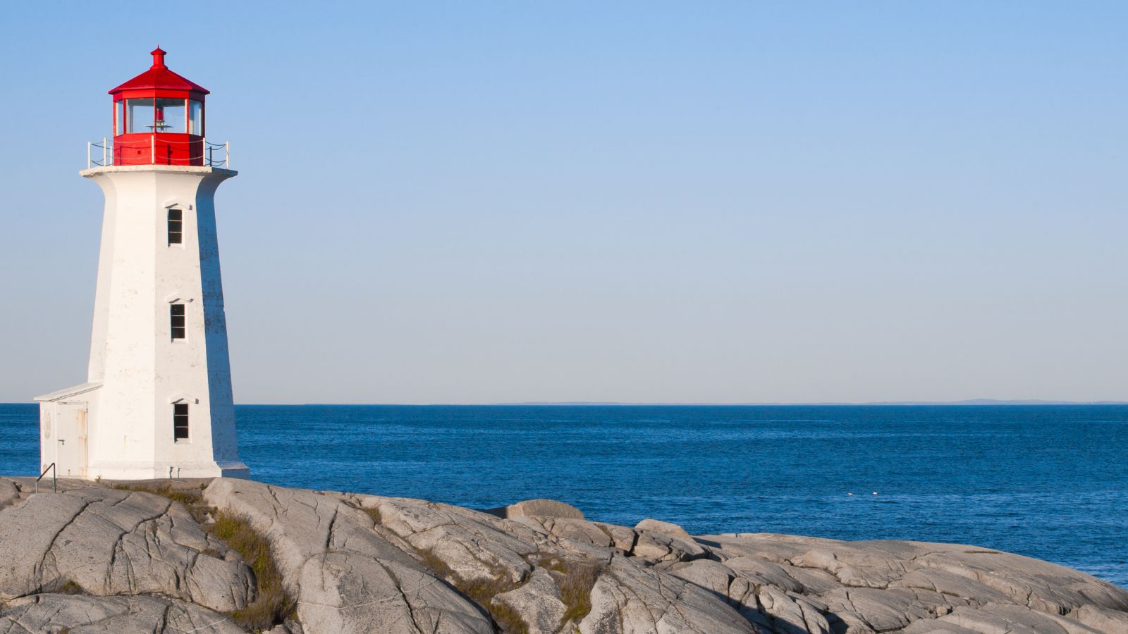 Ein weißer Leuchtturm mit rotem Dach steht auf einem Felsen vor blauem Meer mit blauem Himmel.