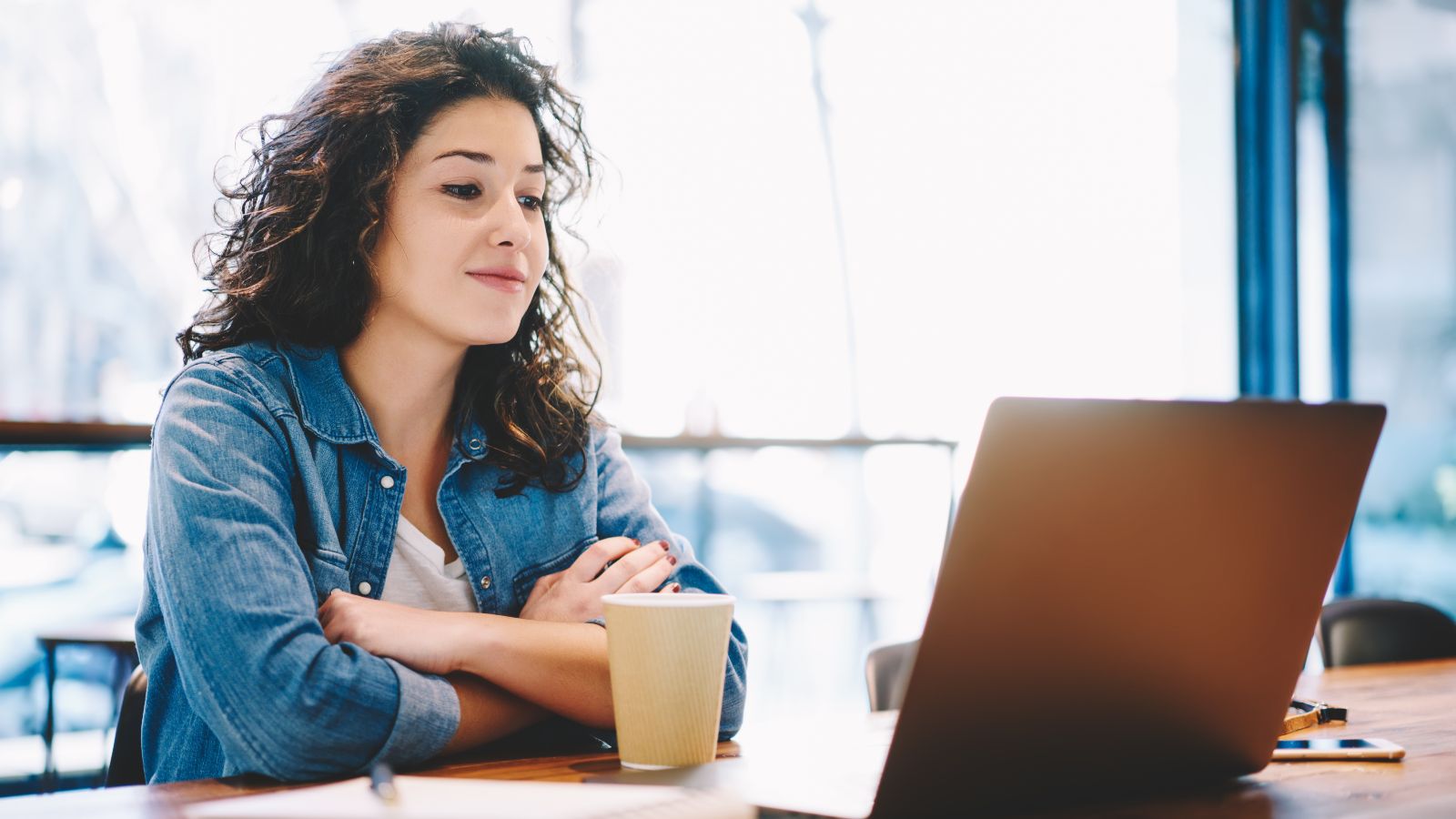 Eine junge Frau sitzt aufmerksam mit Kaffeebecher und Notizblock vor einem aufgeklappten Laptop.